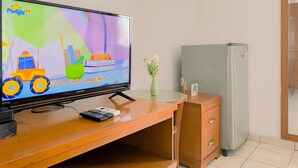 A television on a wooden desk with a remote control, a small vase with flowers, and a refrigerator in a hotel room.
