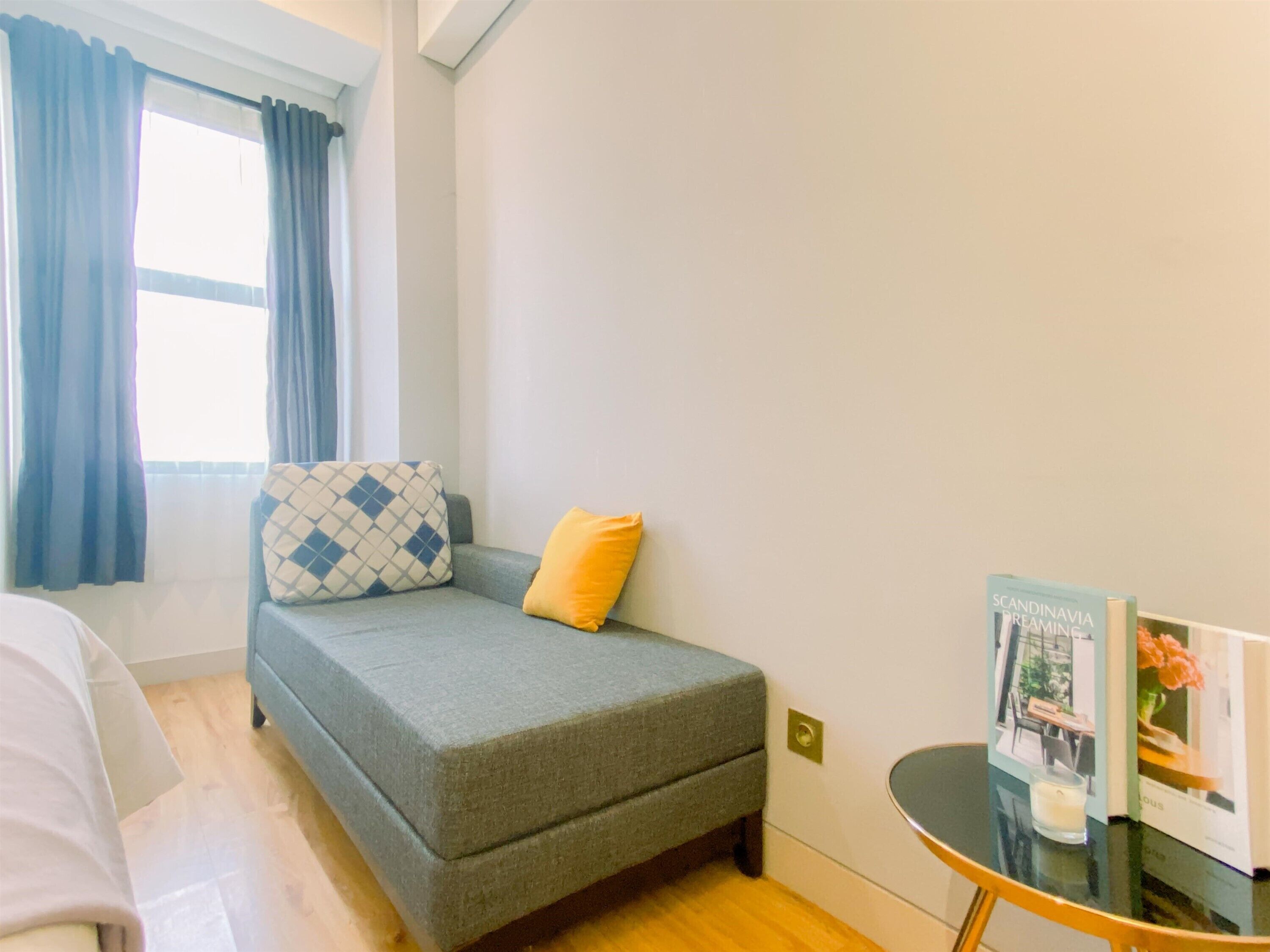 A small, neatly arranged bedroom with a grey sofa bed, a yellow pillow, and a small table with a book and a candle.
