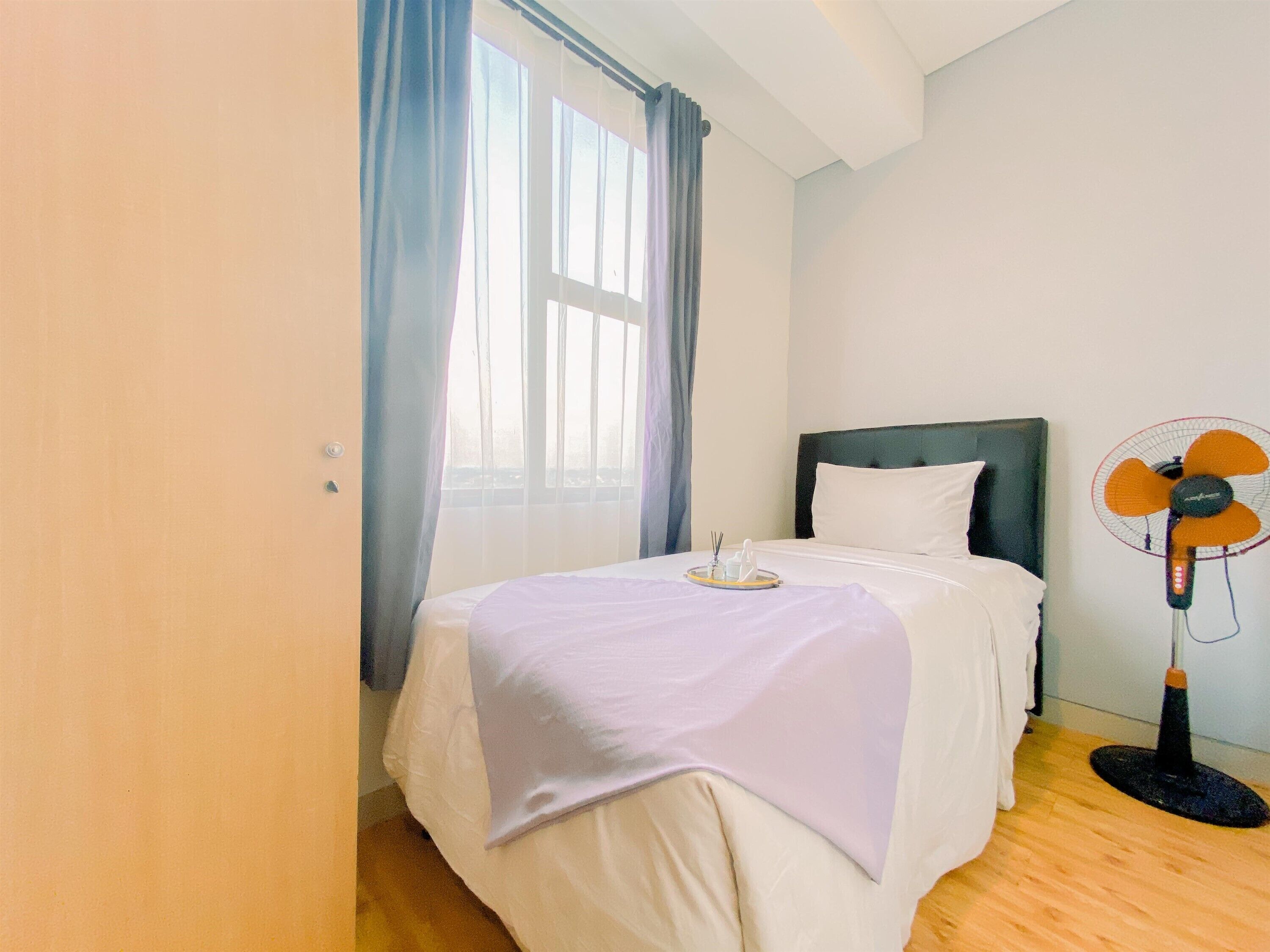 A hotel room with a bed, a bedside table with a lamp, a window with curtains, and a standing fan.