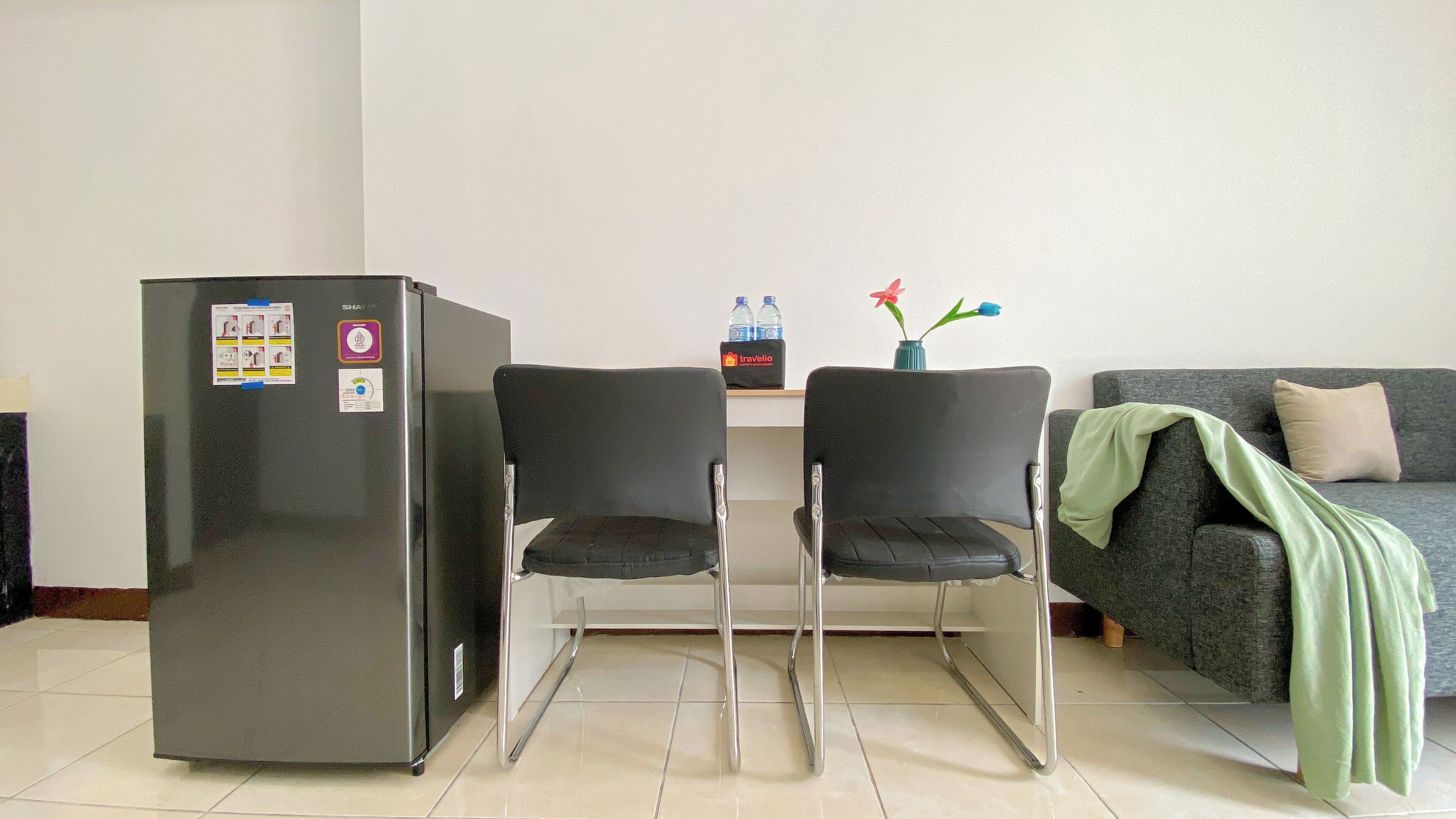 A compact living space with a refrigerator, two chairs, a sofa, a small table with water bottles, and a vase with flowers.