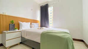 Hotelkamer met een houten hoofdeinde, een nachtkastje met een bord waarop steden staan vermeld, een bed met wit beddengoed, een groene plaid en een raam met gordijnen.