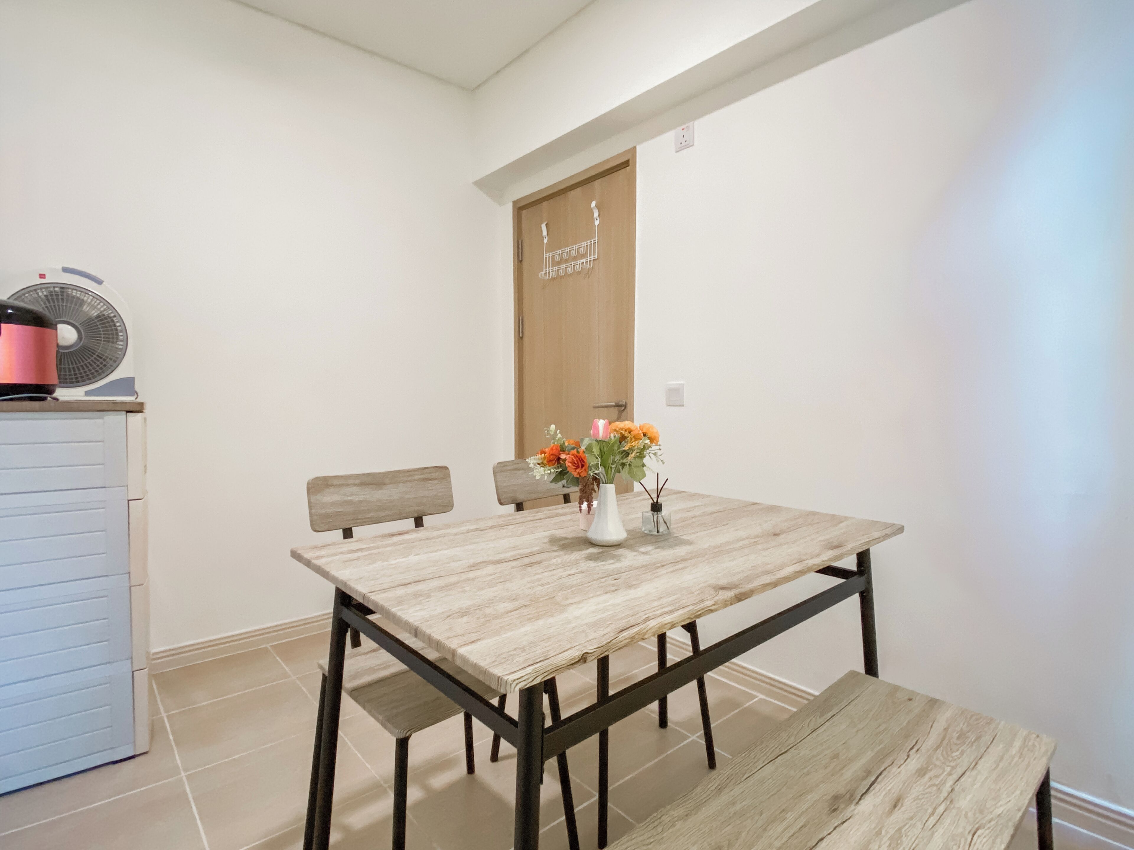 Un comedor con mesa de madera, dos sillas, un banco y un pequeño florero blanco con flores.