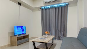 A modern living room with a flat-screen TV, a wooden coffee table, a grey sofa, and a grey curtain. - Simple 2BR at Meikarta Apartment (Cikarang)