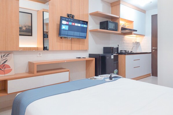 Habitación de hotel pequeña con cama, escritorio, televisor montado en la pared, microondas y una kitchenette.