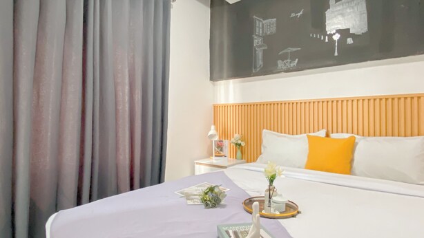 A hotel room with a bed, a nightstand, a wall-mounted artwork, and a window with curtains. - Simply Look 1Br At Boutique Kemayoran Apartment (Jakarta)