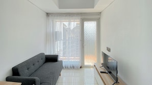 A modern living room with a grey sofa, a flat-screen TV on a wooden stand, and a sliding glass door leading to an outdoor area. - Simply Look 1Br At Boutique Kemayoran Apartment (Jakarta)