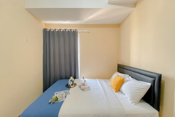 A hotel room with a bed, a blue bedspread, white and yellow pillows, a small table with a flower arrangement, and a curtain. - Comfort Living 1Br Boutique Kemayoran Apartment (Jakarta)