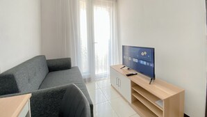 A modern living room with a grey sofa, a TV on a wooden stand, and a large window with sheer curtains. - Comfort Living 1Br Boutique Kemayoran Apartment (Jakarta)