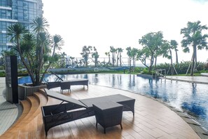 A poolside area with lounge chairs and a small table, surrounded by palm trees and a modern building in the background. - Modern And Tidy 2Br At Tanglin Supermall Mansion Apartment (Surabaya)