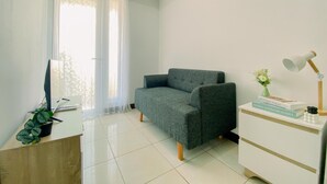 Ein modernes Wohnzimmer mit einem grauen Sofa, einem kleinen Schreibtisch mit einem Laptop, einer Topfpflanze und einer Lampe.