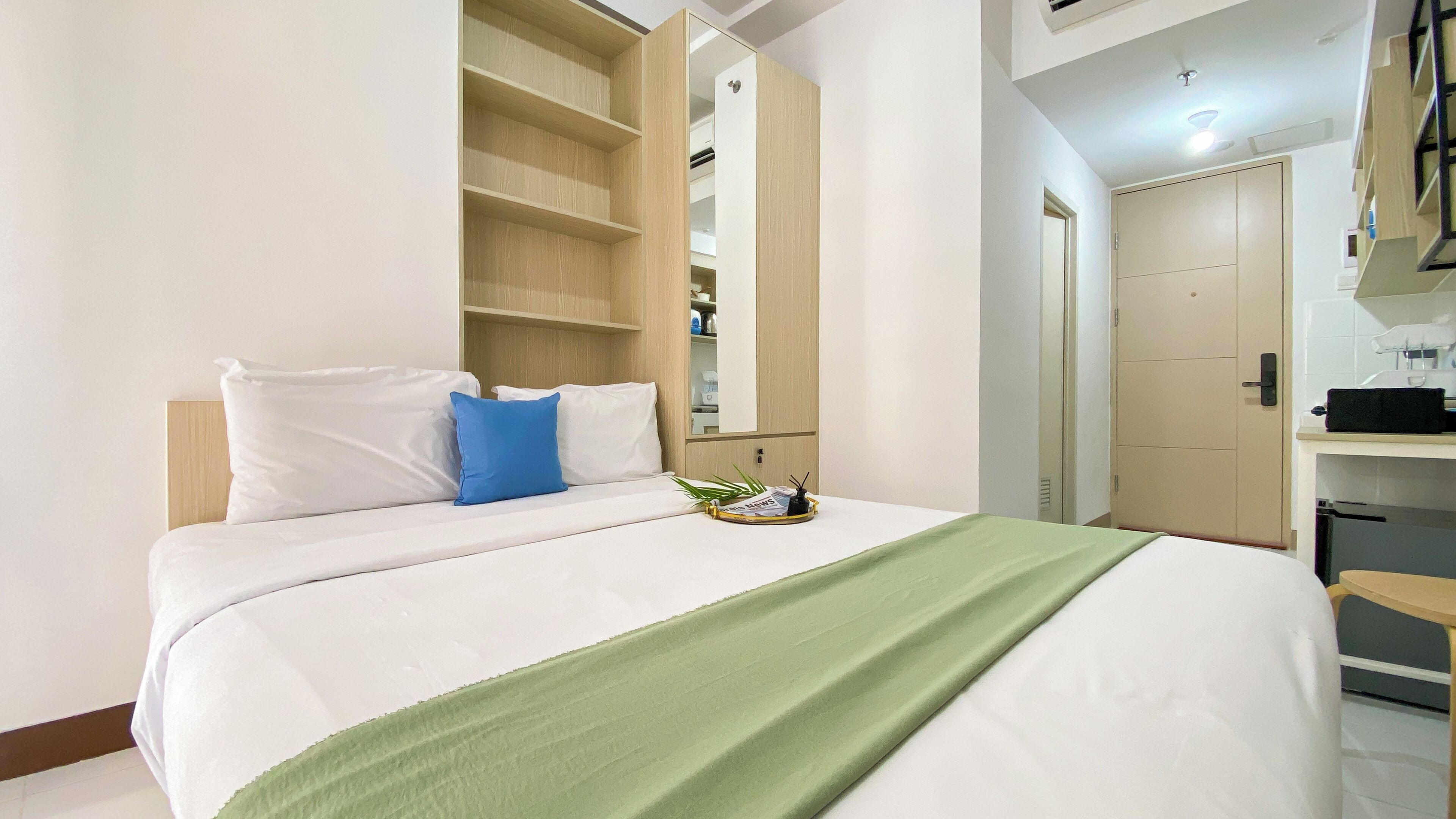 A neatly made bed with white and blue pillows, a green throw blanket, and a tray with a newspaper and a pen on the nightstand.