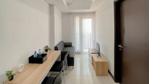 Een moderne, goed verlichte kamer met een houten bureau, een zwarte bank en een televisie op een houten standaard. Op het bureau staat een kleine plant en aan de rechterkant een deur.