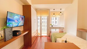 Une chambre d’hôtel moderne avec une télévision sur le côté gauche, un bureau en bois, un lit recouvert d’une couverture verte et un balcon offrant une vue sur la ville.
