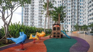 Un espace de jeux pour enfants, doté d’équipements colorés et d’un toboggan, entouré d’un jardin aménagé et avec un grand immeuble d’habitation en arrière-plan.
