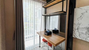 A modern hotel room with a wooden desk, two red bar stools, a large mirror, and a view of greenery through sheer curtains.