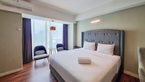 Quarto de hotel com uma cama grande, duas cadeiras, uma mesinha e uma janela com cortinas.