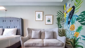 Quarto com cama e cabeceira estofada cinza, sofá cinza com almofadas, piso de madeira e parede com obras de arte de tema tropical.