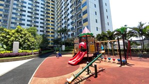 A playground with a red slide and swings, surrounded by a red and black rubberized surface, with tall residential buildings in the background. - Best Place And Restful Studio At Apartment Springlake Summarecon Bekasi (Bekasi)