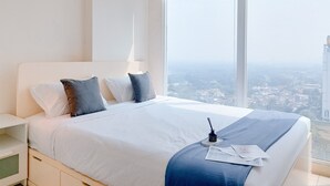 Habitación de hotel con una cama grande, ropa de cama blanca, una mesita de noche, una lámpara y vistas a un paisaje urbano a través de amplias ventanas.