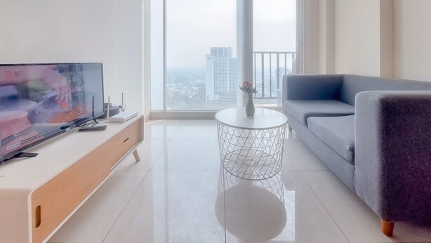 Una sala de estar moderna con un televisor grande, un sofá gris, una mesa de centro redonda y vistas a un paisaje urbano a través de amplios ventanales.