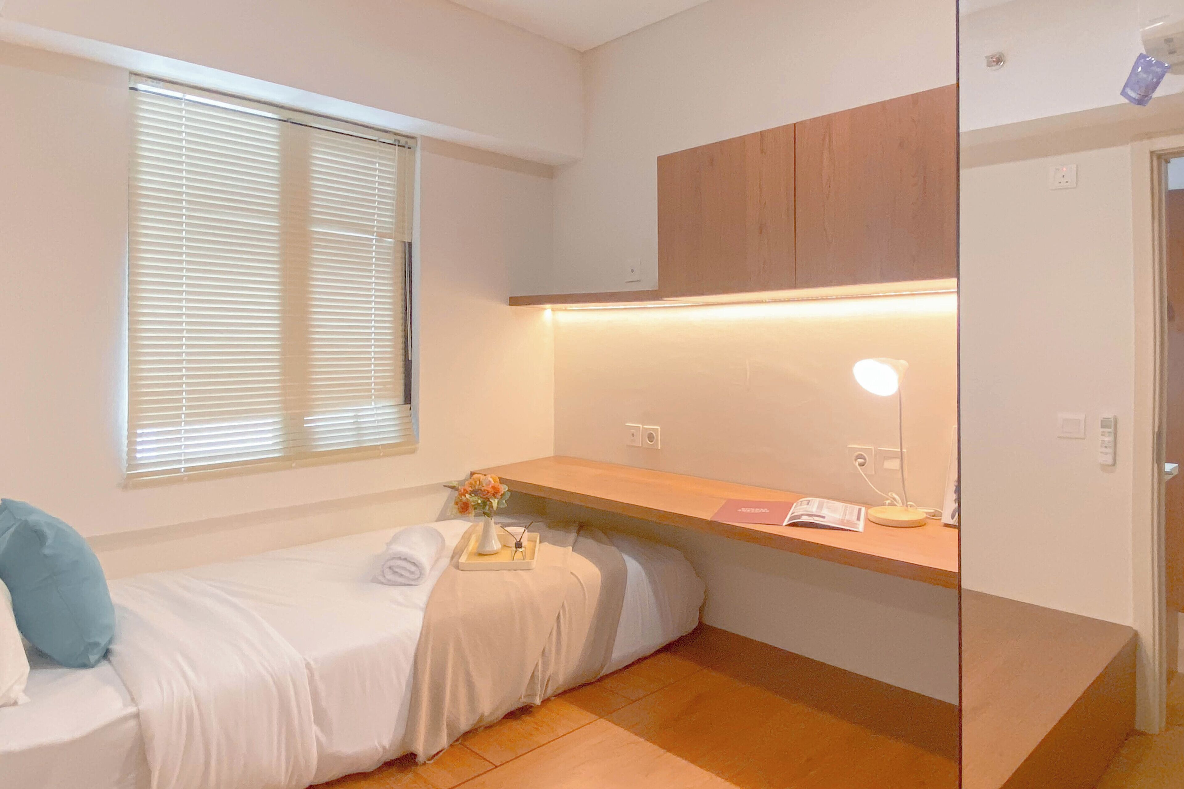 Ein kleines, ordentlich eingerichtetes Schlafzimmer mit einem Einzelbett, einem Nachttisch mit Lampe, einem Fenster mit Jalousien und einem Holzschrank über dem Bett.