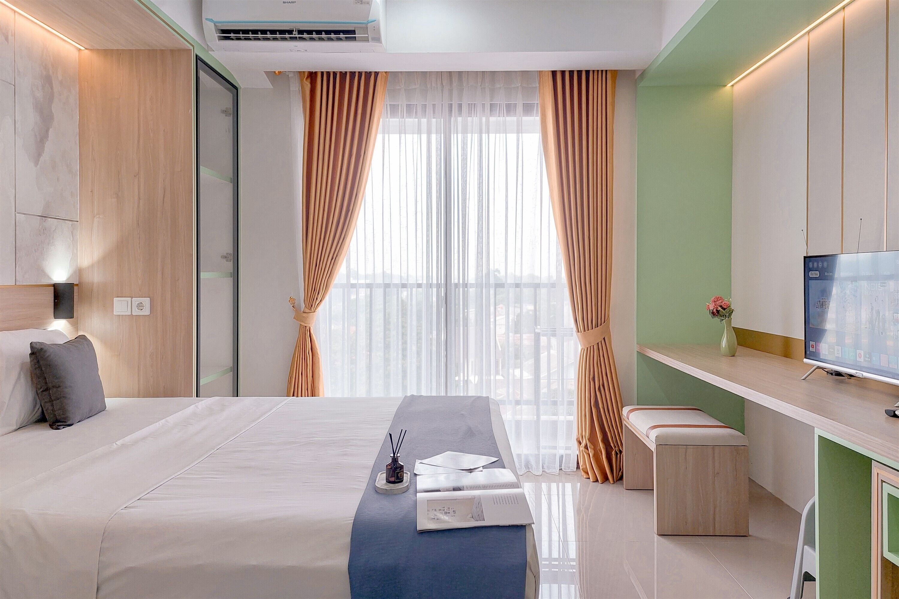 A modern hotel room with a bed, bedside table, TV, and a view of the outdoors through sheer curtains.