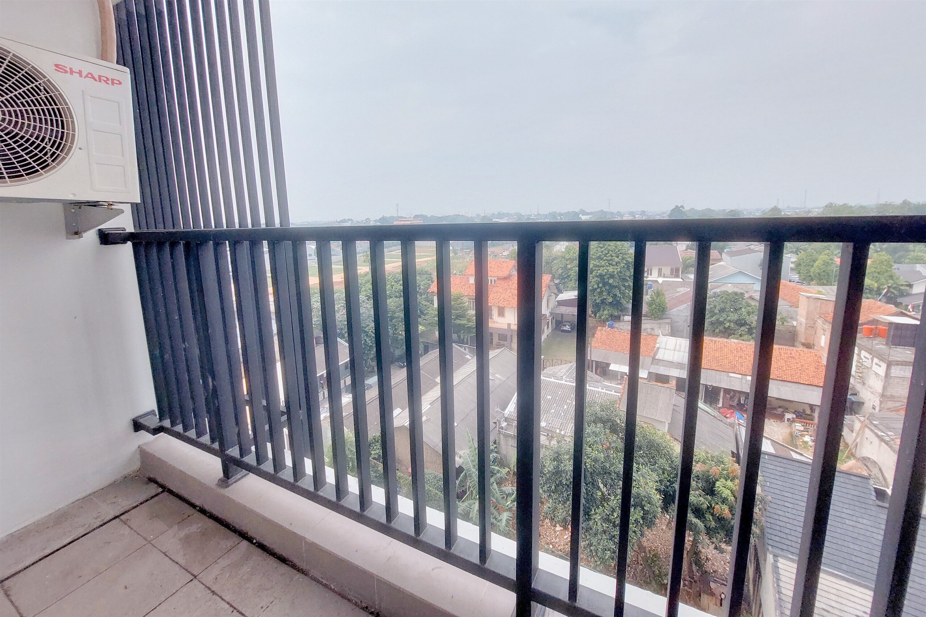 A balcony with a black metal railing, a Sharp air conditioner mounted on the wall, and a view of the neighborhood below.