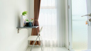 A small room with a white chair, a shelf with books and a plant, a door with a textured glass panel, and sheer curtains. - Best Deal And Restful Studio At Amazana Serpong Apartment (South Tangerang)