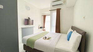 A neatly arranged hotel room with a bed, a television mounted on the wall, a window with curtains, and an air conditioning unit. - Best Deal And Restful Studio At Amazana Serpong Apartment (South Tangerang)