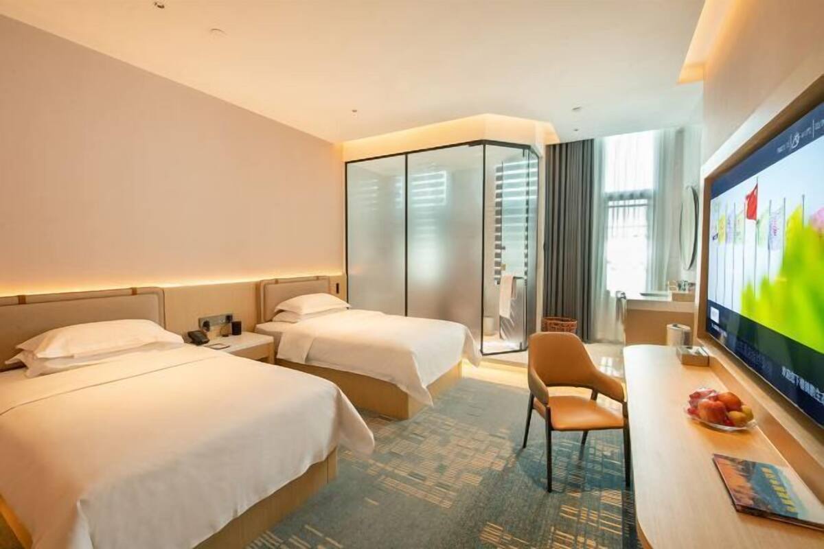 Ein Hotelzimmer mit zwei Betten, einem Schreibtisch, einem Sessel, einem Fernseher und einer Glaskabine.
