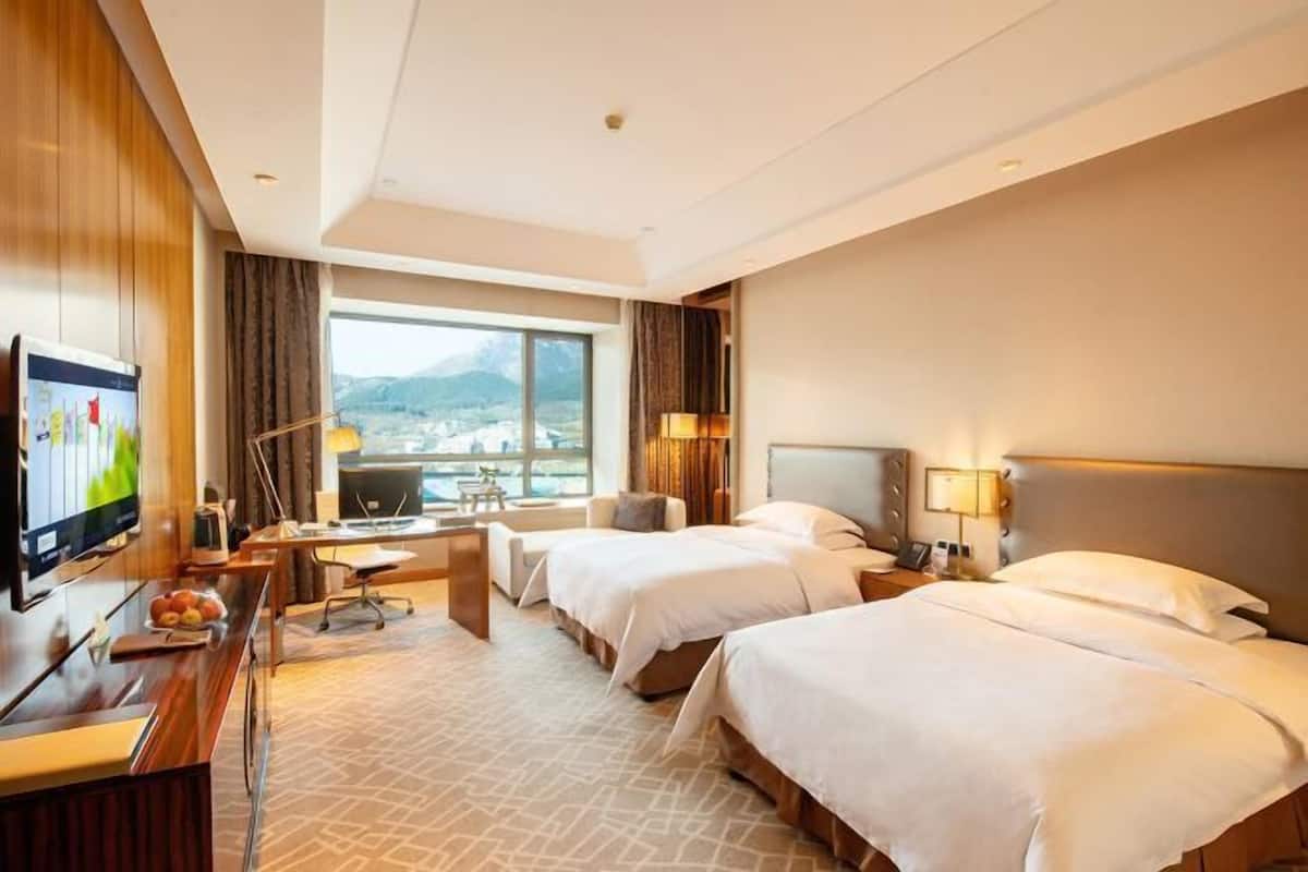 Ein Hotelzimmer mit zwei Betten, einem Schreibtisch mit Computer, einem Fernseher und Blick auf die Stadt.
