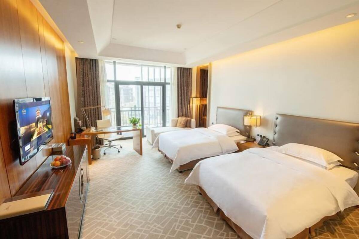 Ein Hotelzimmer mit zwei Betten, einem Schreibtisch mit Fernseher, einem Fenster mit Vorhängen und einer Pflanze.