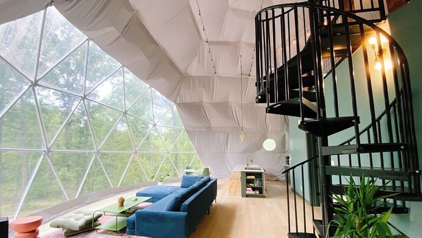 Un intérieur moderne avec un plafond aux motifs géométriques, un canapé bleu, un escalier en colimaçon et une grande fenêtre donnant sur un paysage verdoyant.