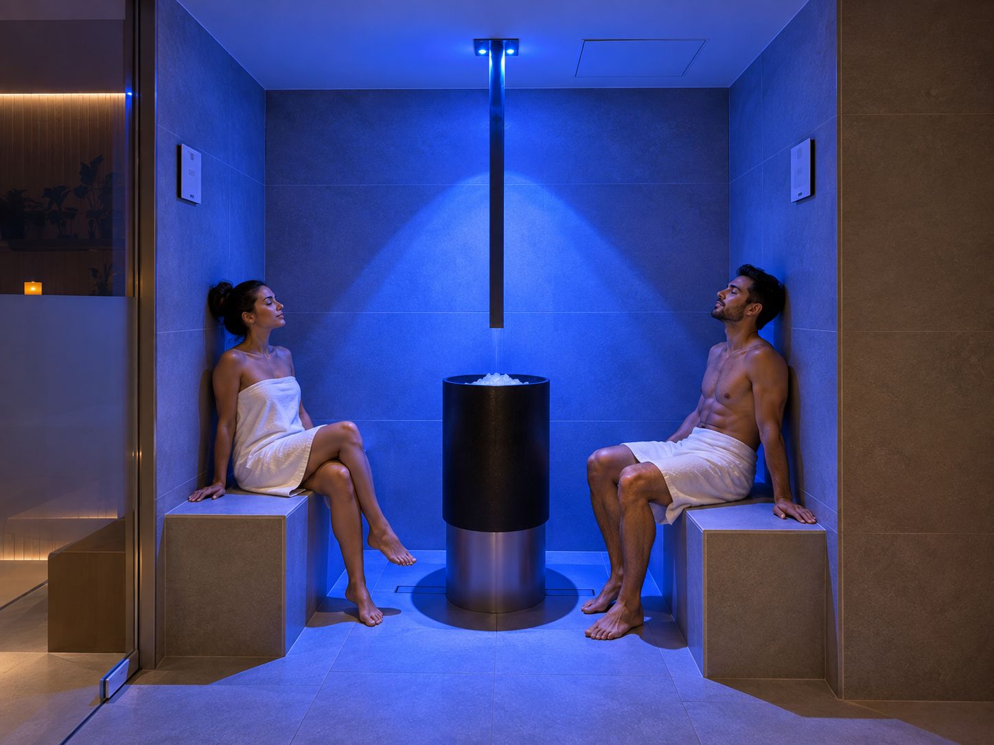 Dos personas sentadas en una sauna con un elemento calefactor central, rodeadas de paredes azuladas y un suelo de baldosas.