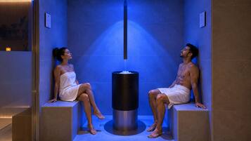Dos personas sentadas en una sauna con un elemento calefactor central, rodeadas de paredes azuladas y un suelo de baldosas.