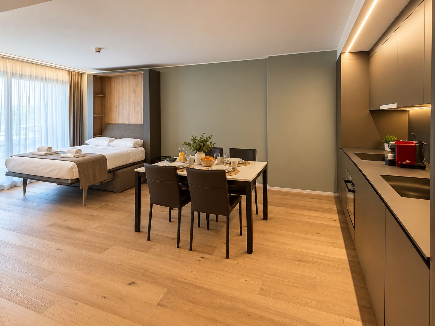 Habitación de hotel moderna con cama, una mesa de comedor con sillas, zona de cocina y un amplio ventanal con cortinas.
