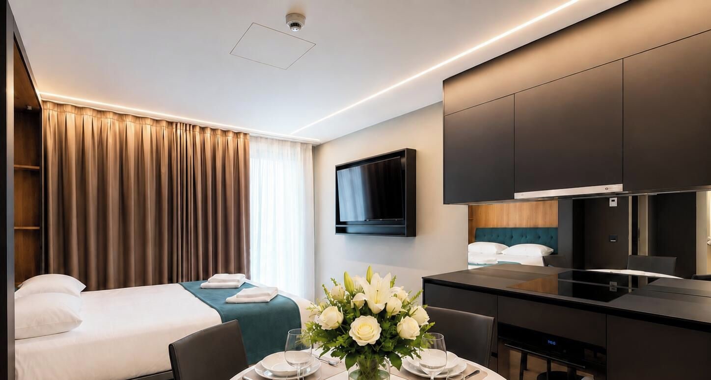 A hotel room with a bed, a desk, a flat-screen TV, and a dining area with a vase of flowers.