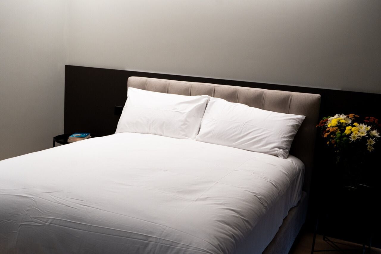 A neatly made bed with white linens and a headboard, a bedside table with a book, and a vase with flowers.