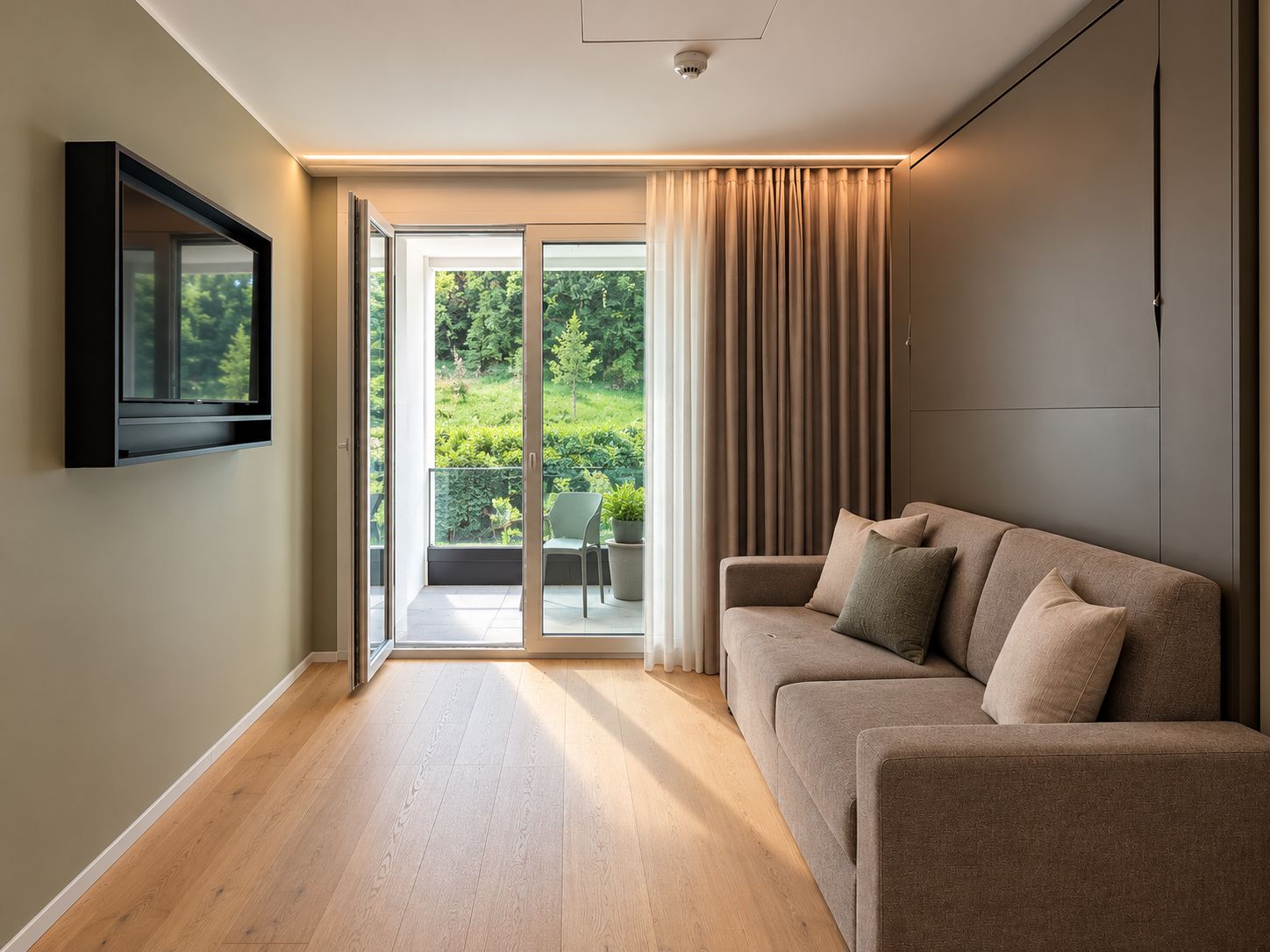A modern living room with a sofa, a flat-screen TV mounted on the wall, and a sliding glass door leading to a balcony with a view of greenery.
