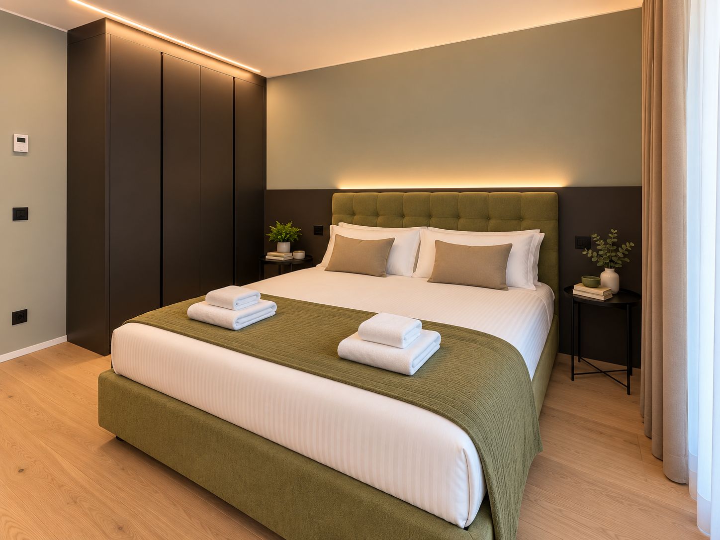 Une chambre d’hôtel moderne avec un grand lit, une armoire en bois foncé, des tables de chevet ornées de plantes et un banc recouvert d’un tissu vert.
