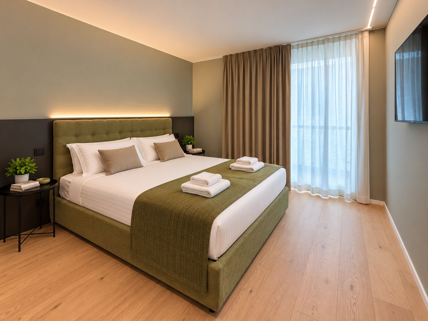 A modern hotel room with a large bed, a bedside table with a plant, and a TV mounted on the wall.
