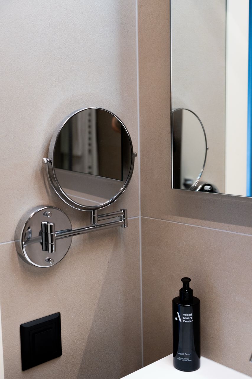 A bathroom with a round mirror mounted on the wall, a black bottle of liquid soap, and a black electrical outlet.
