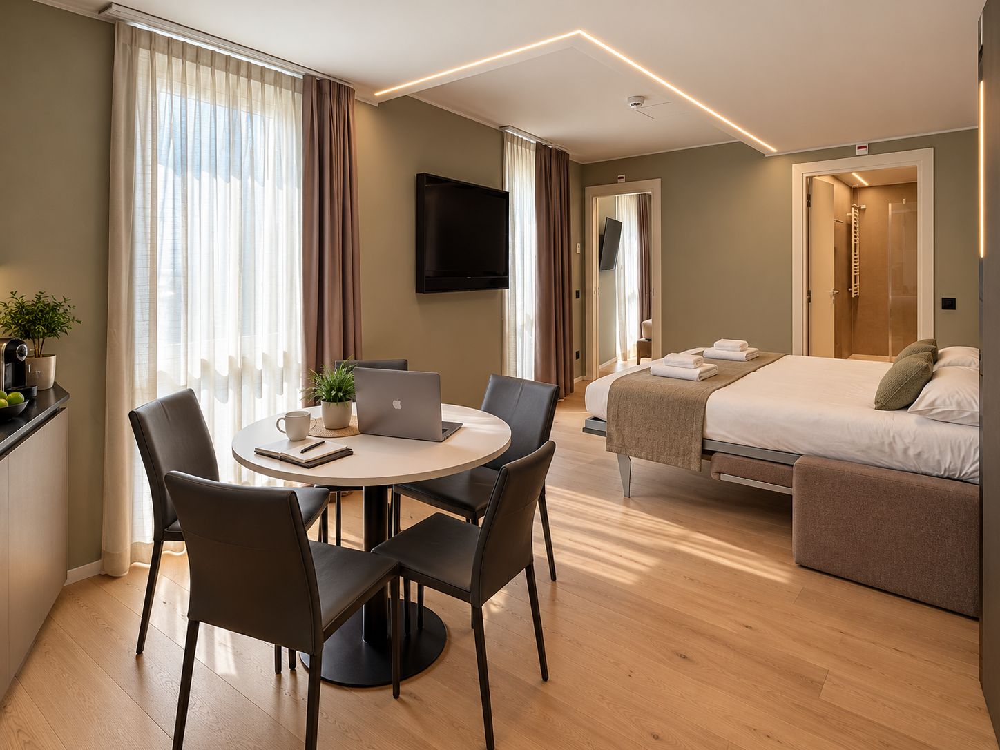 Quarto de hotel com cama, escrivaninha com laptop, cadeira, televisão e banheiro visível através de uma porta aberta.