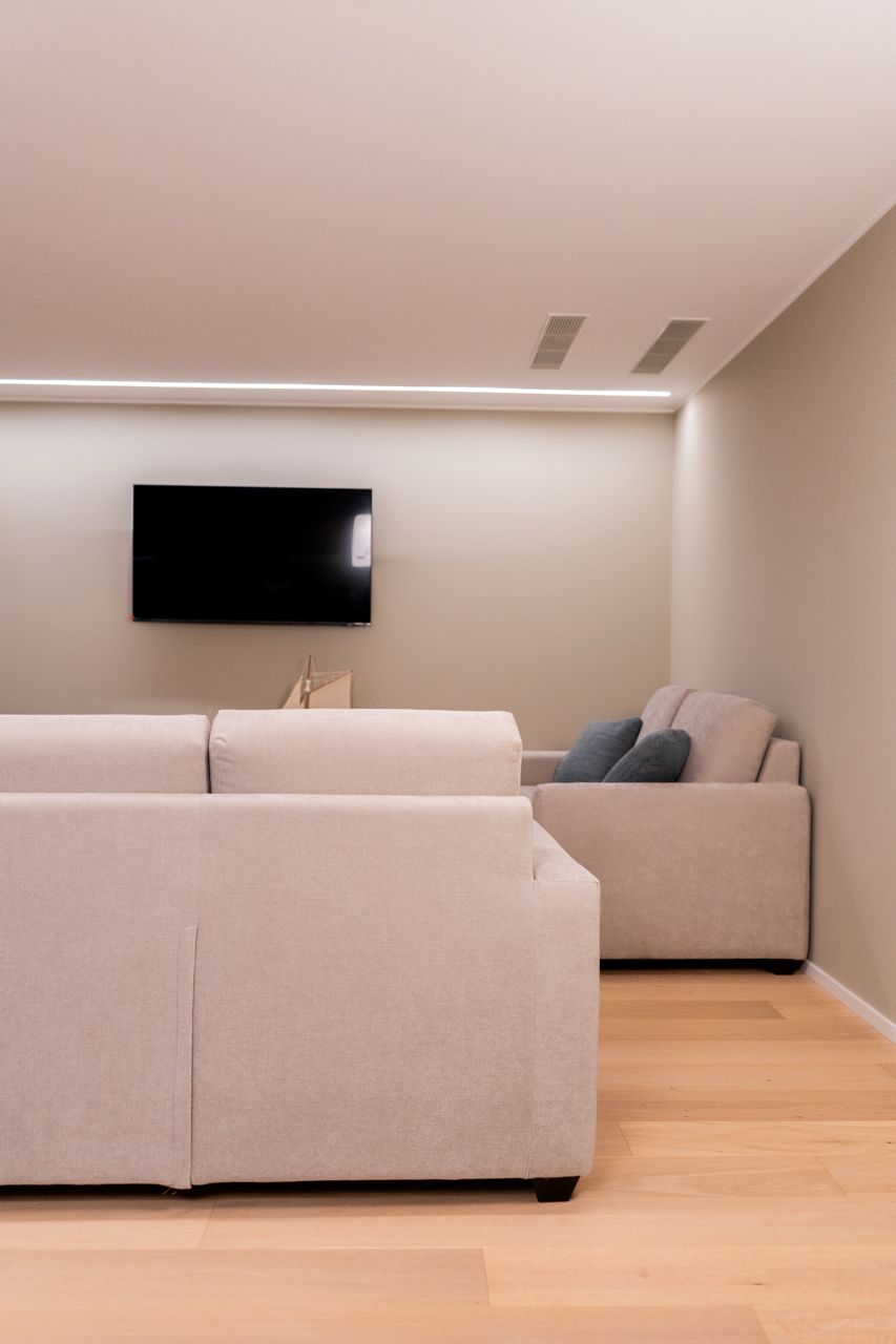 A modern living room with a large flat-screen TV mounted on the wall, a beige sofa, and a smaller armchair.