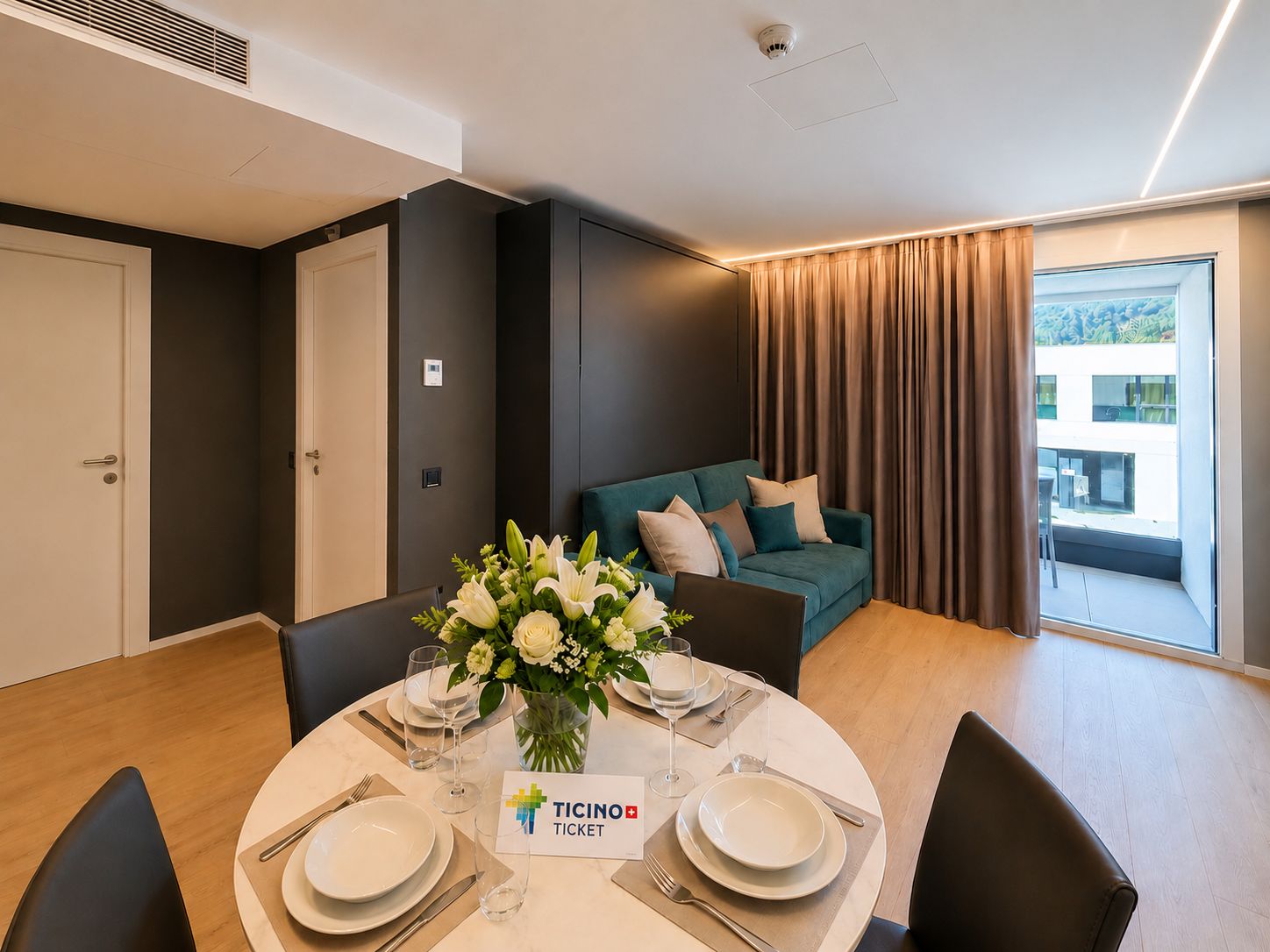 Een moderne eethoek met een ronde tafel gedekt voor twee personen, een bank op de achtergrond en uitzicht buiten door een groot raam.
