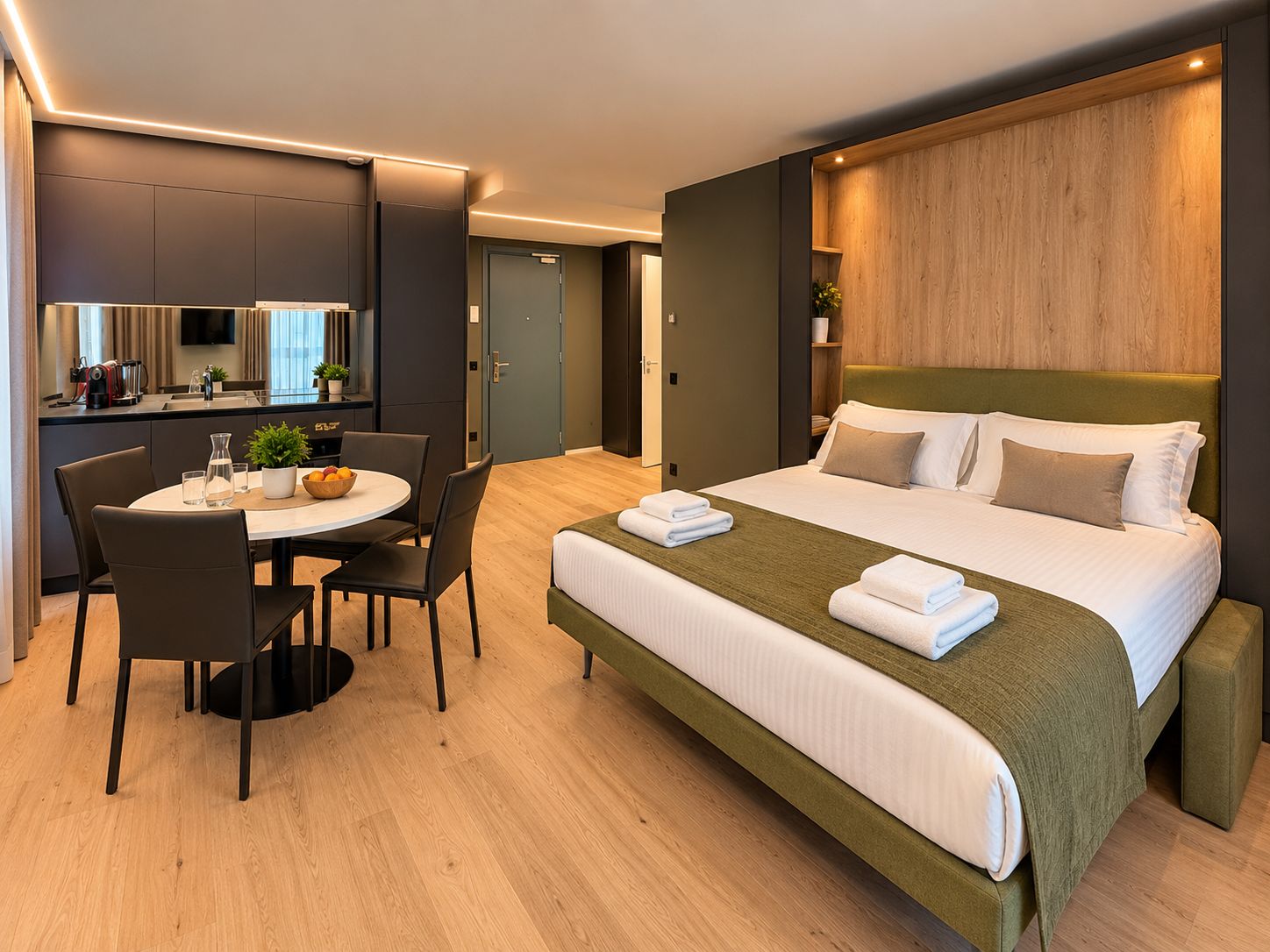 A modern hotel room with a large bed, a dining table with chairs, a kitchen area, and a television.