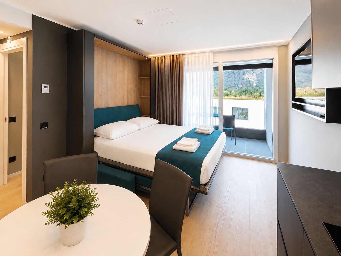 Une chambre d’hôtel moderne équipée d’un lit, d’un bureau, d’une chaise, d’une plante en pot et d’un balcon avec vue.