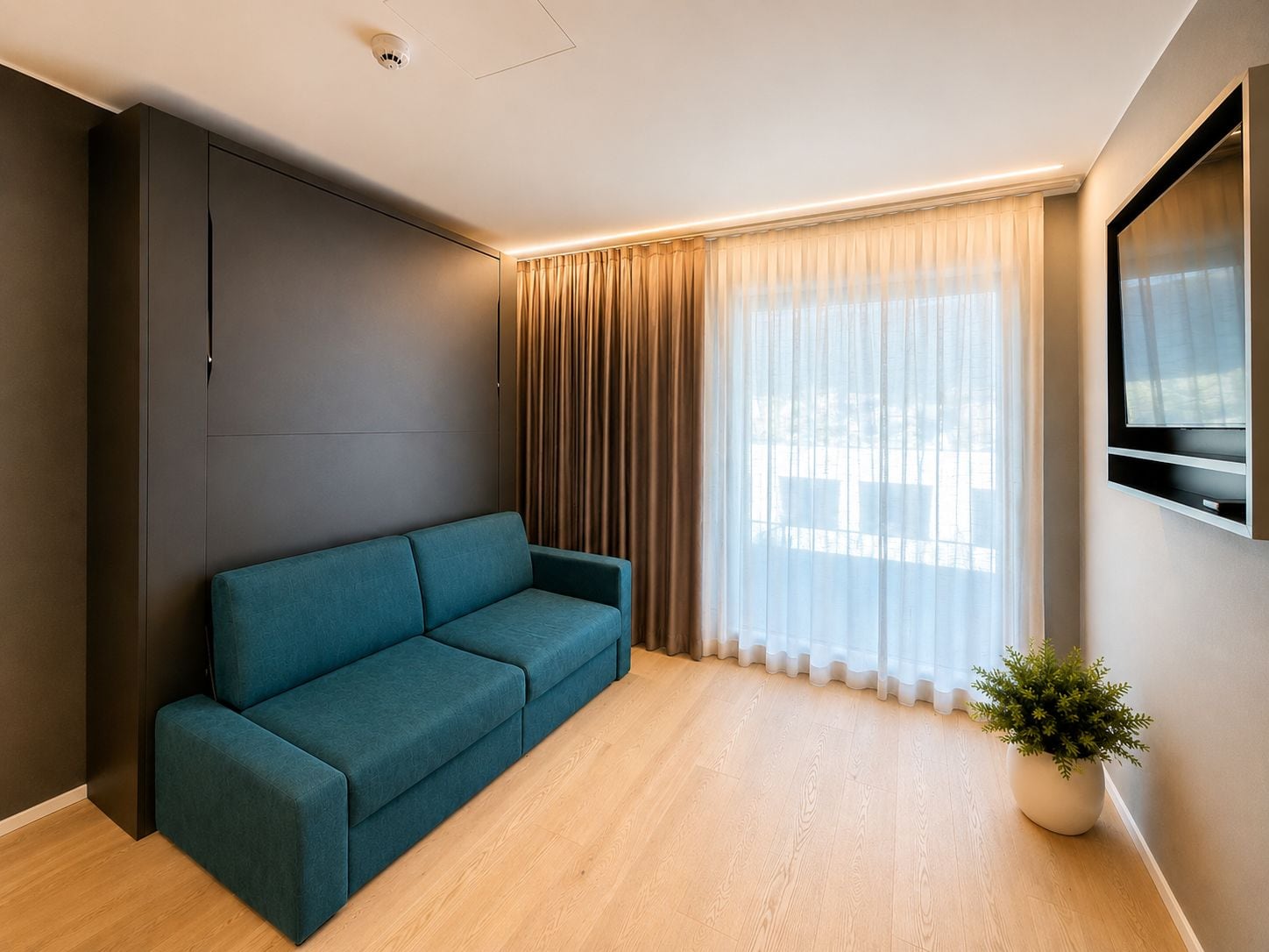 Ein modernes Wohnzimmer mit einem türkisfarbenen Sofa, einem großen Fenster mit transparenten Vorhängen, einem Wand-Fernseher und einer Topfpflanze.