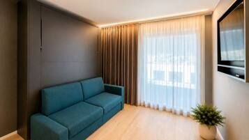 A modern living room with a teal sofa, a large window with sheer curtains, a mounted TV, and a potted plant.