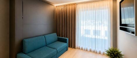 Ein modernes Wohnzimmer mit einem türkisfarbenen Sofa, einem großen Fenster mit transparenten Vorhängen, einem Wand-Fernseher und einer Topfpflanze.